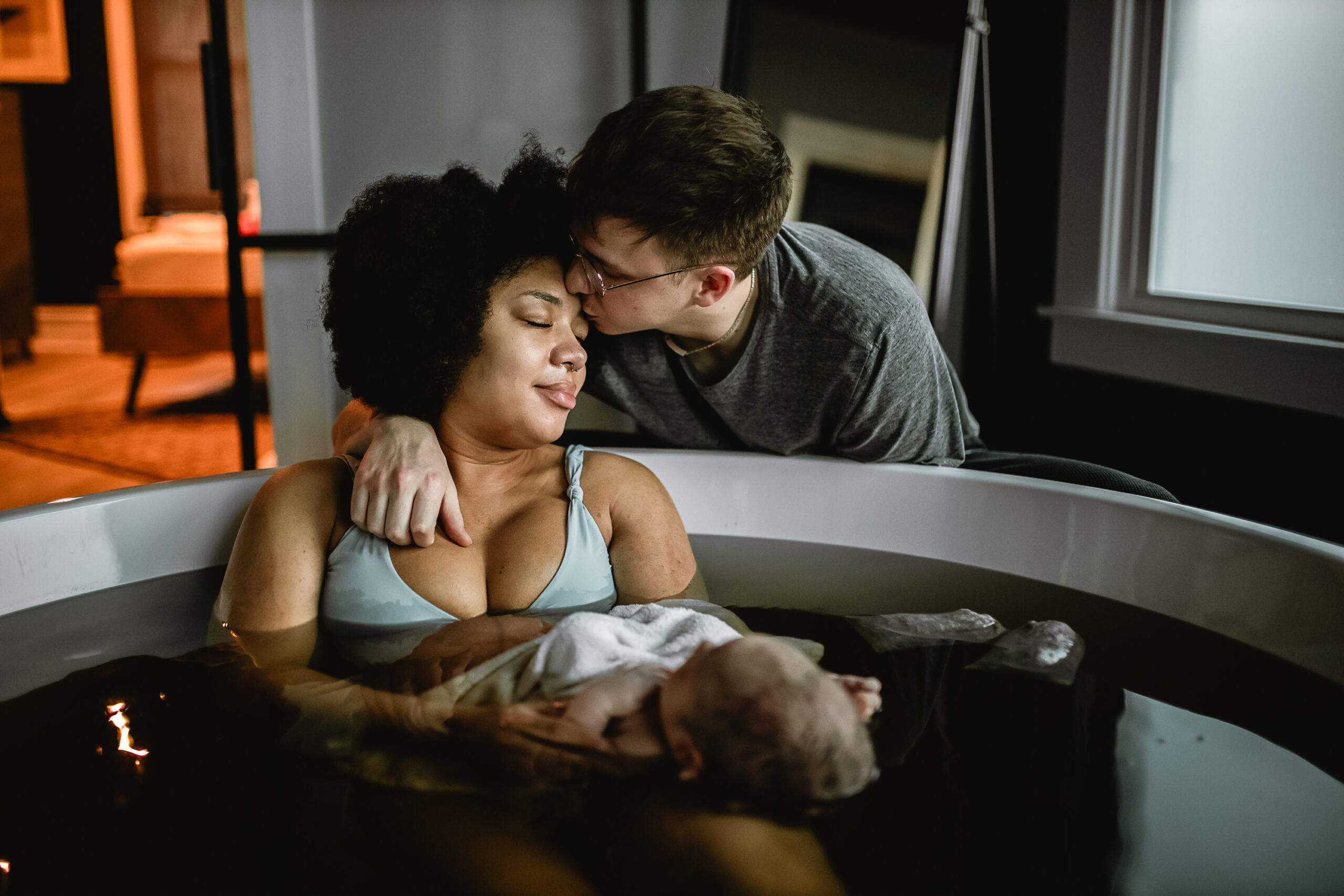 Mother holding her newborn during an herbal bath at a home birth in Fort Worth, with her husband leaning close as they both look down at their baby. Documentary birth photography in DFW.