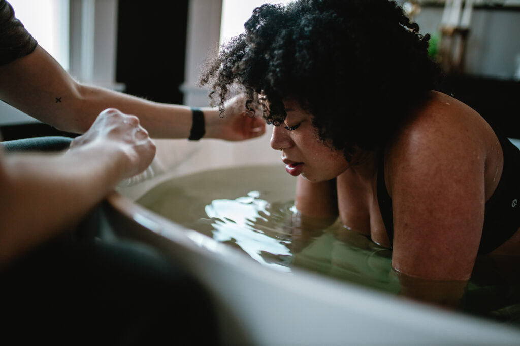 First-time mother focused in active labor, leaning forward in the birth tub as she breathes through a contraction