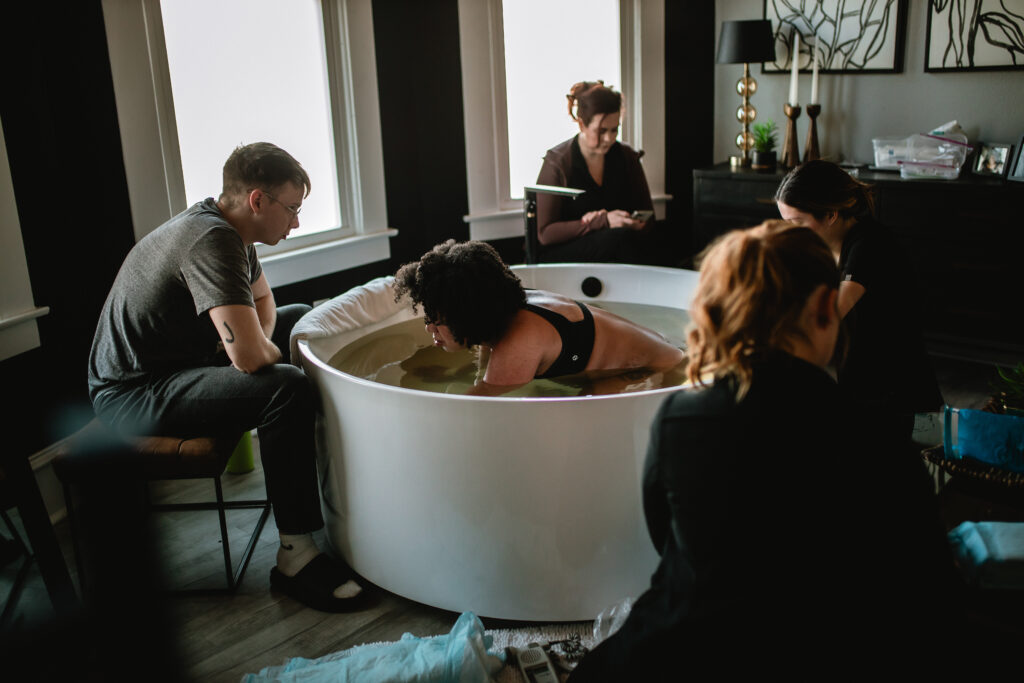 Wide view of the birth room — mother laboring in the tub surrounded by her husband, midwives, and support team