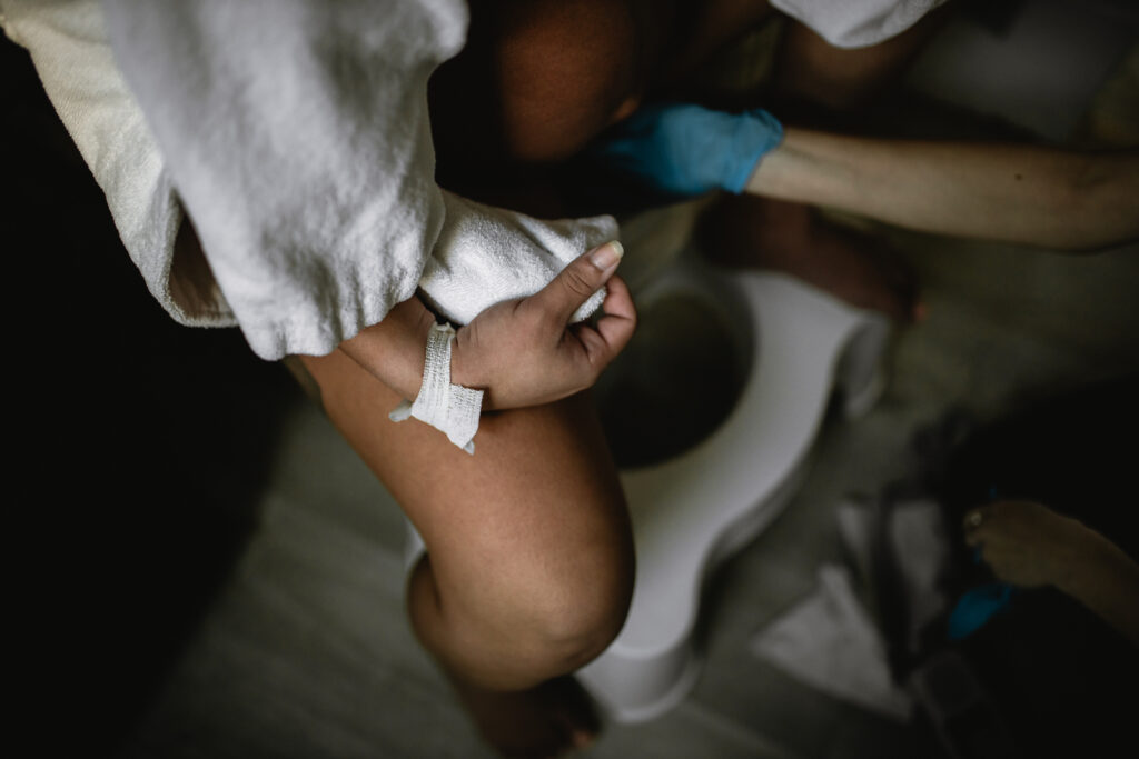 Close-up of the mother’s legs and hands as the midwife supports her while she labors on the birth stool