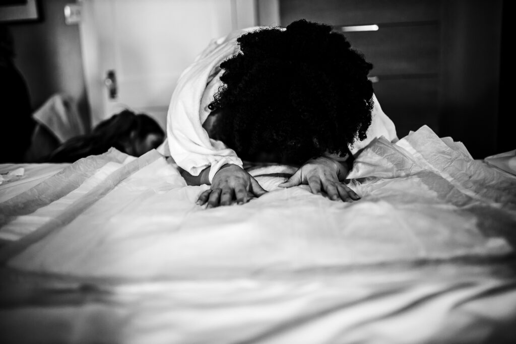 Black and white photo of the mother leaning forward on the bed, gripping the birth sheets during labor