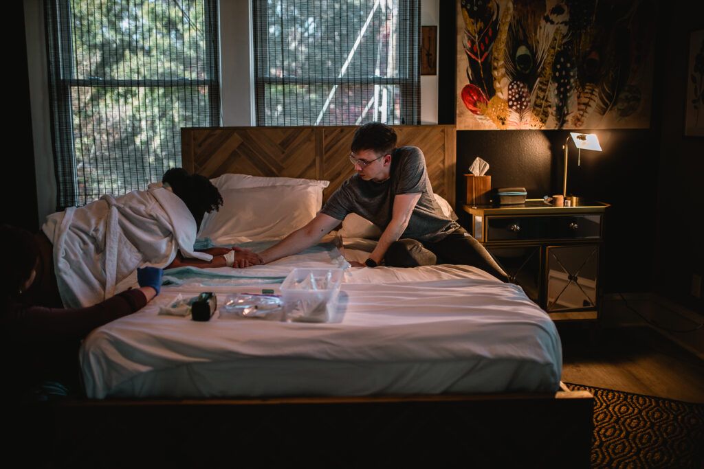 Husband kneeling beside his wife on the bed, holding her hands and grounding her through a powerful contraction