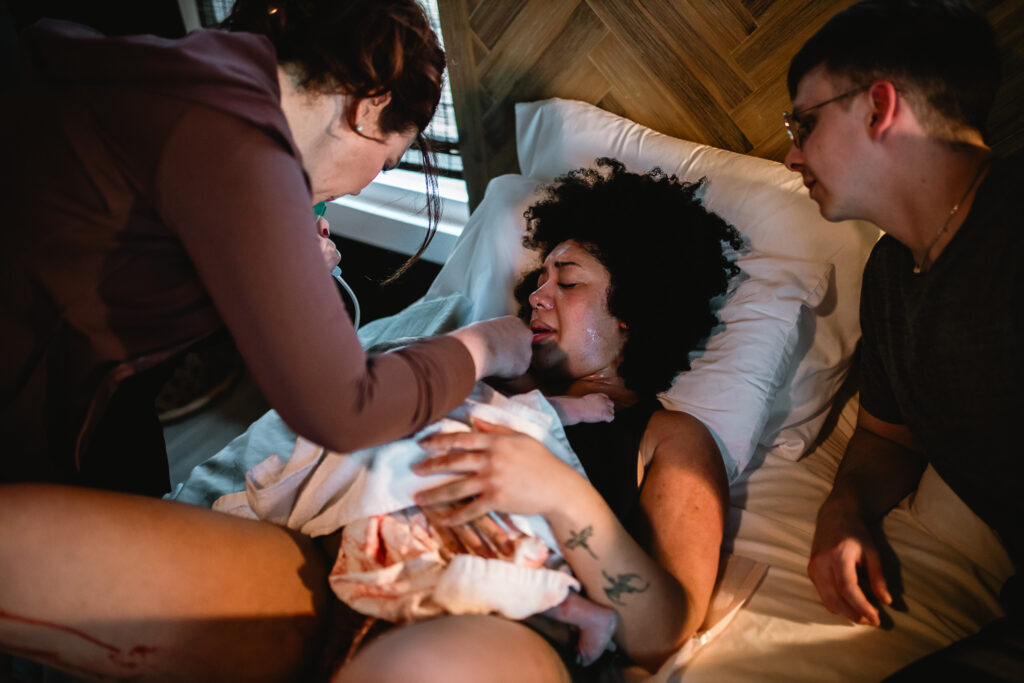 Mother resting on the bed with her newborn on her chest while her birth team offers support