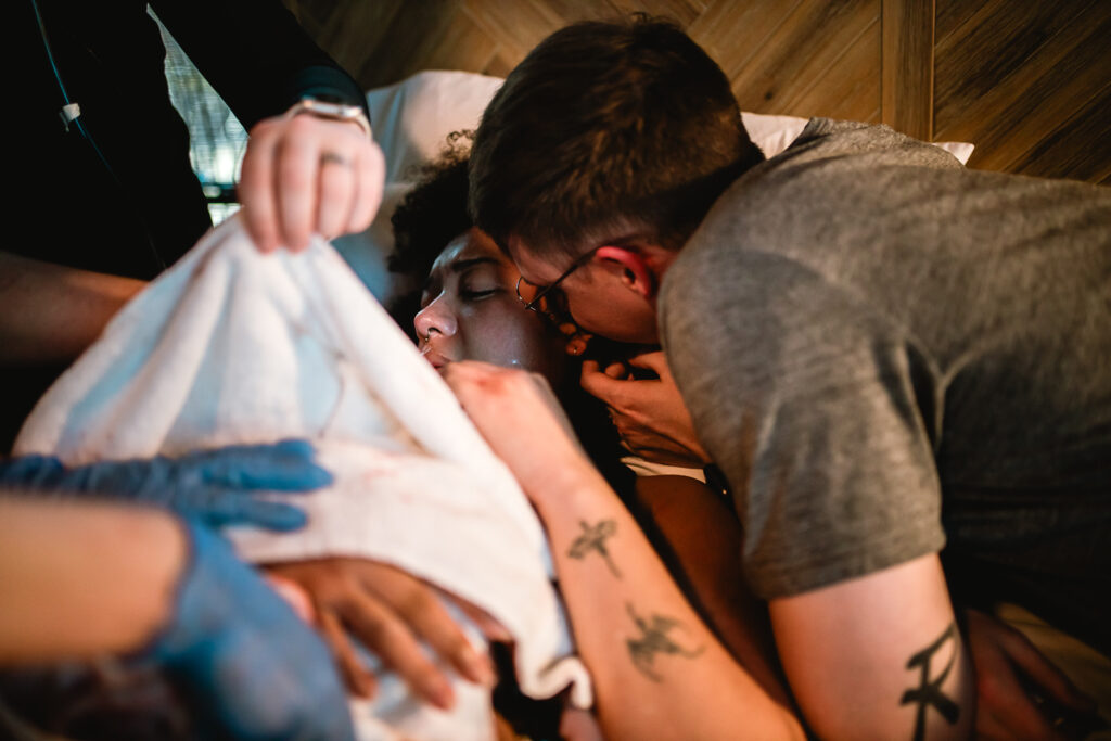 Mother lying back on the bed while her husband cuddles in close, taking in the first quiet minutes as a family