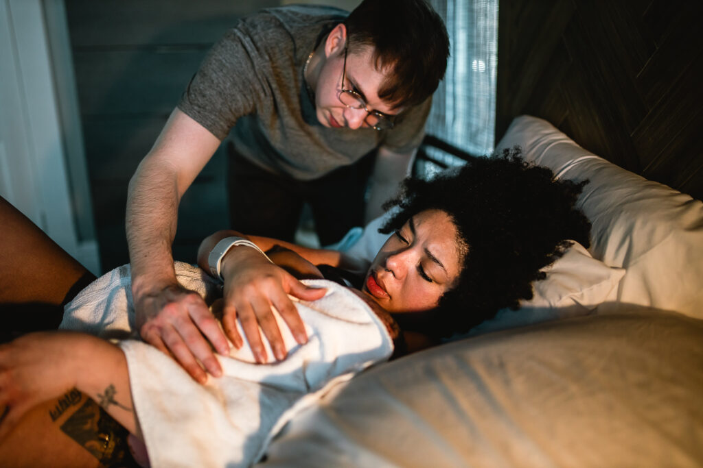 Father gently rubbing the mother’s shoulder while she rests in bed with their newborn