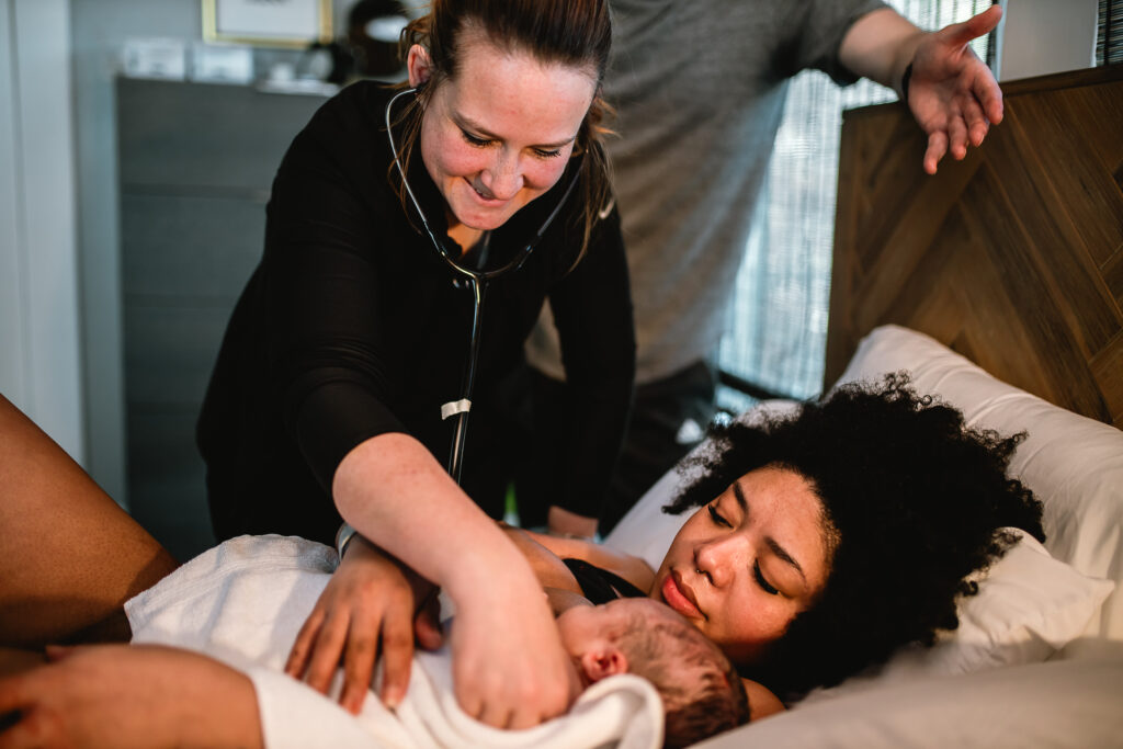 Midwife carefully checking the newborn while the mother rests on pillows, watching over her baby