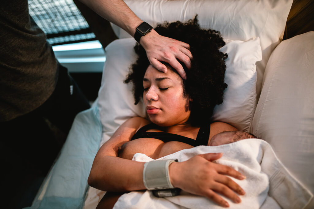 Large photo — Midwife checking the newborn’s vitals while the mother lies in bed, cradling her baby in the first quiet postpartum moments