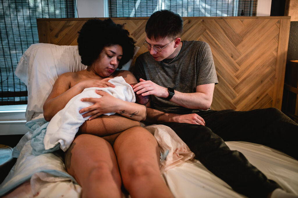 Mother holding her newborn on her chest while father gently touches the baby, both parents resting together in bed after birth