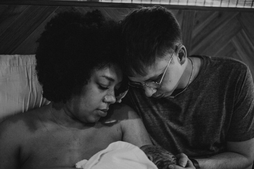 Black and white photo of parents resting with their newborn, foreheads touching as they hold their baby close