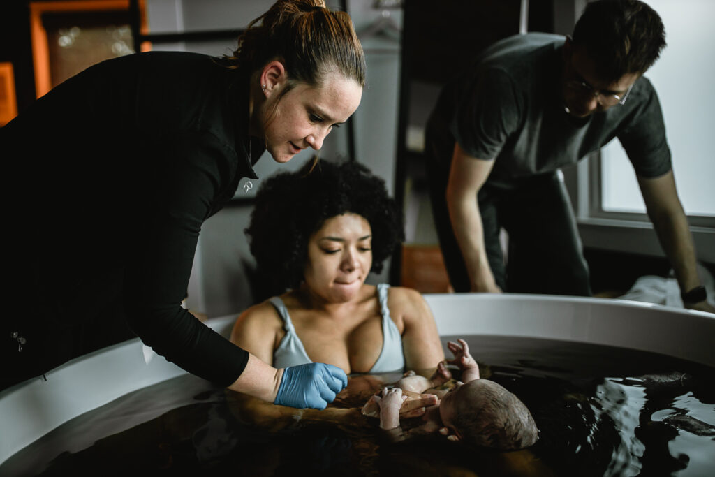 Midwife assisting during newborn herbal bath while parents support their baby in the birth tub