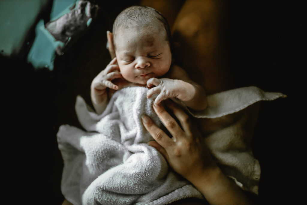 Newborn wrapped in a towel being lifted from the herbal bath, tiny hands tucked near their face