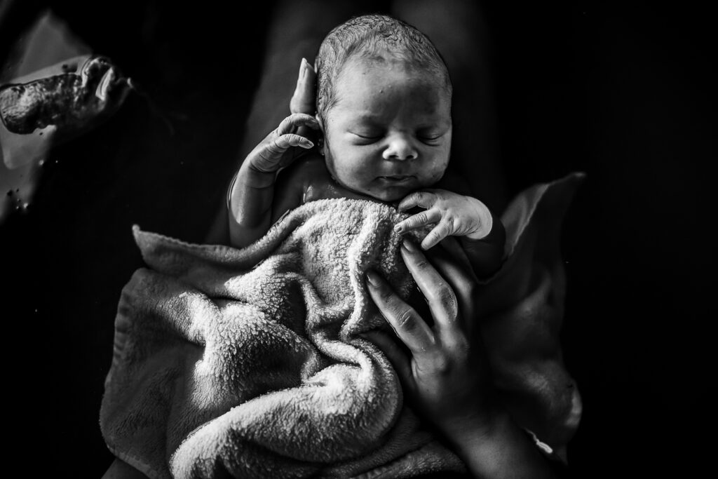 Black and white close-up of newborn’s face wrapped in towel, tiny hand beside cheek