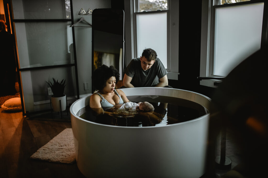 Mother resting in the birth tub holding her newborn while her partner sits beside her