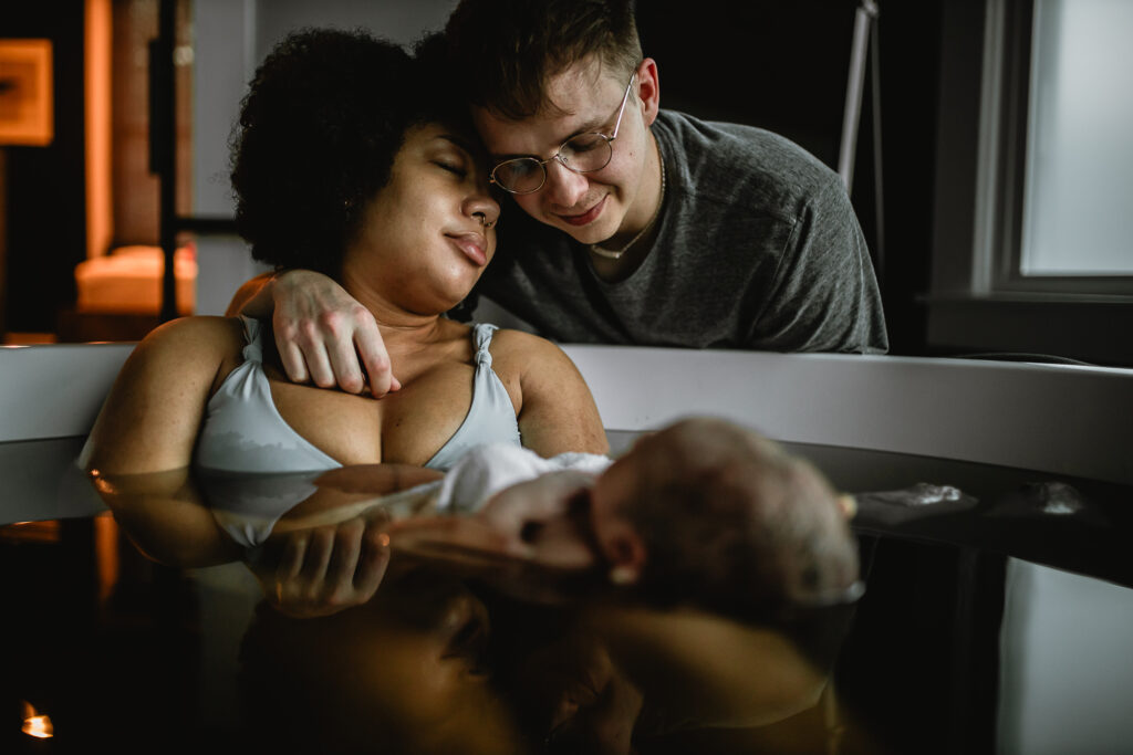 Mother leaning back in the tub while father wraps his arm around her and looks at their baby