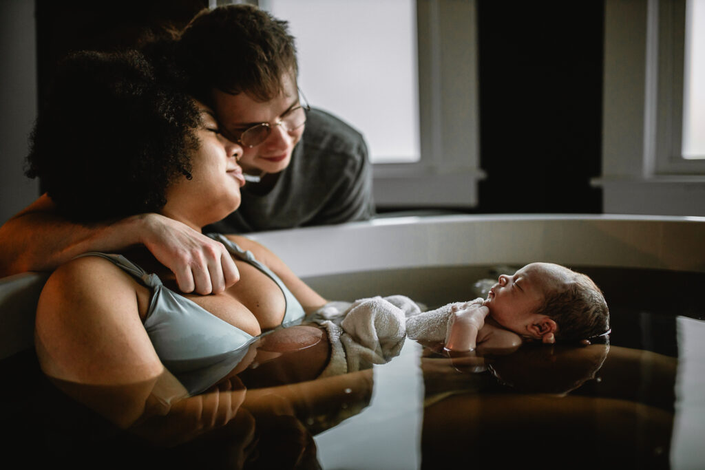 Parents holding newborn together in the tub, both smiling softly