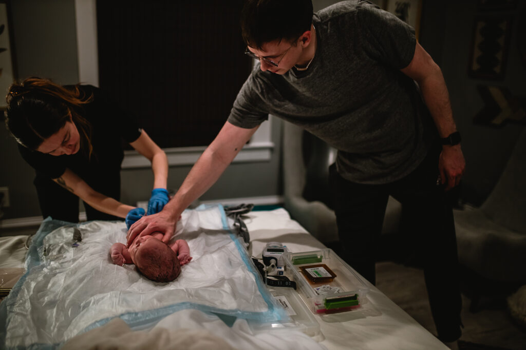 Father reaches out to touch newborn while midwife finishes exam