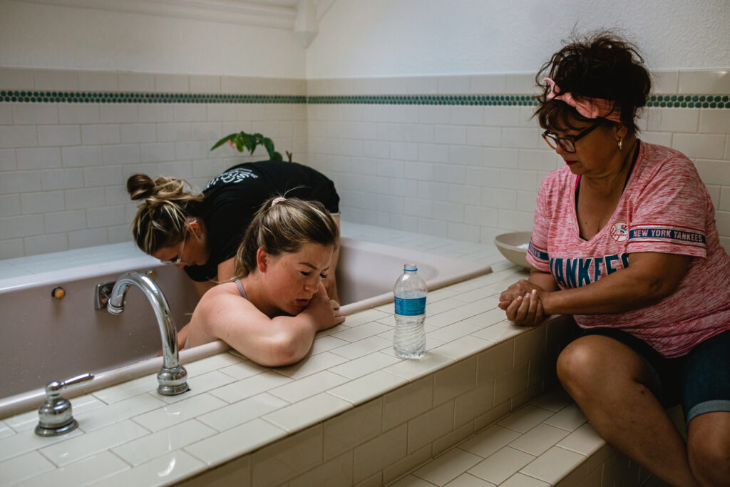 Mother laboring at home birth while leaning over bathtub with family nearby