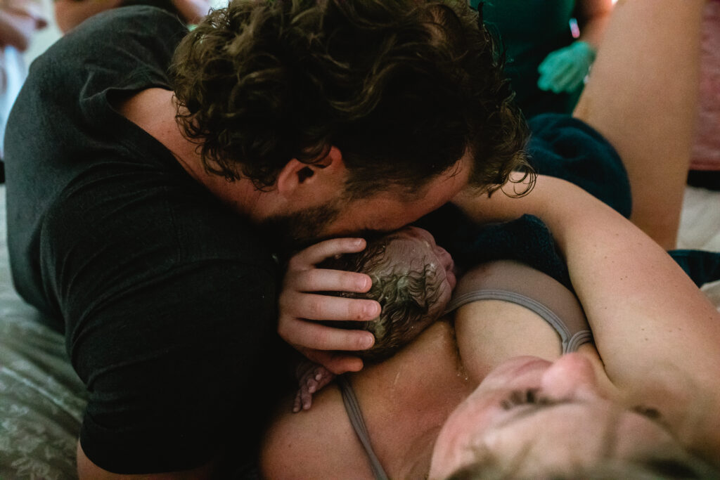 Father kissing mother’s forehead while newborn rests on her chest during first moments after home birth