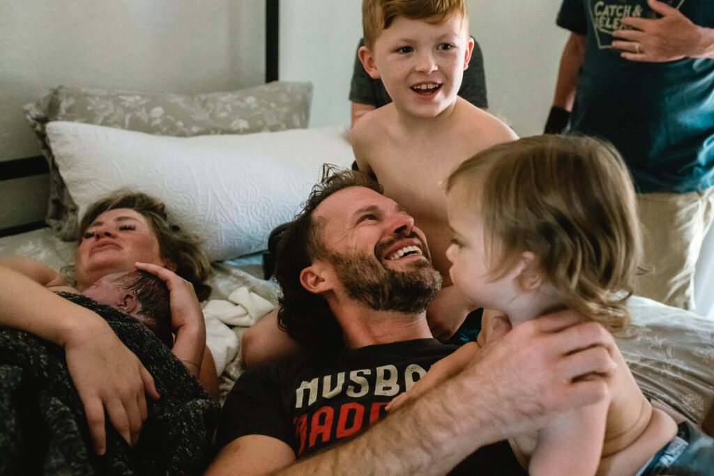 Family laughing together on bed as big brothers gather around their newborn sibling after home birth