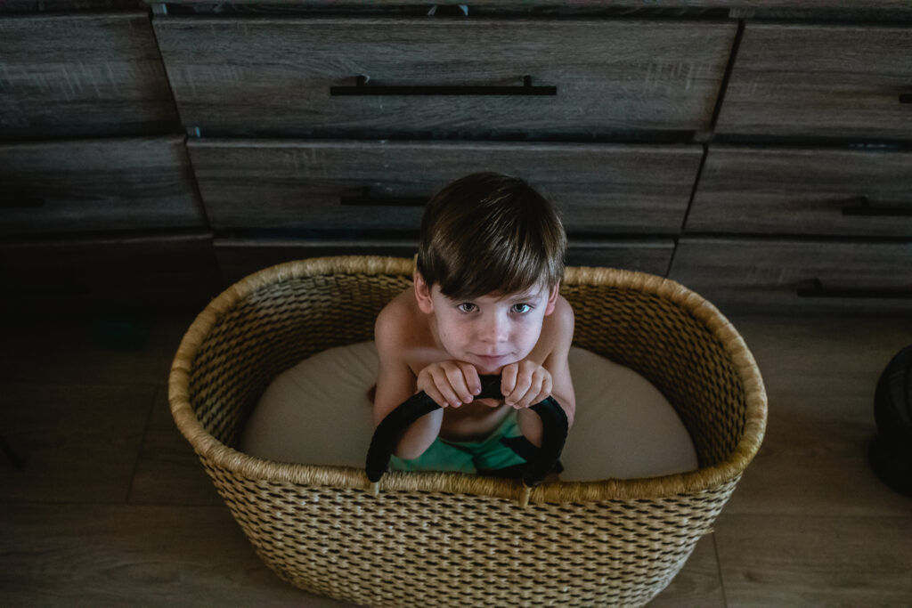 Toddler sitting playfully inside empty Moses basket during quiet moments after baby’s home birth