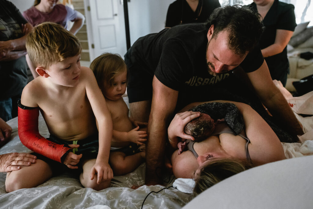 Older brothers sitting beside parents while newborn rests skin-to-skin on mother’s chest