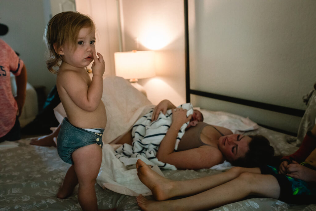 Mother resting in bed holding newborn while older sibling watches gently nearby