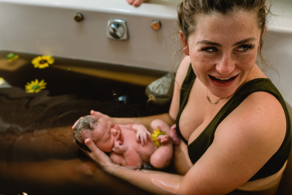 Newborn resting peacefully in herbal bath, tiny hands curled beside face, surrounded by sunflowers