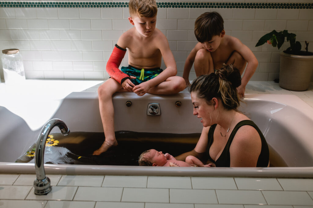 Big brothers gathered around tub watching newborn in herbal bath during home birth postpartum care