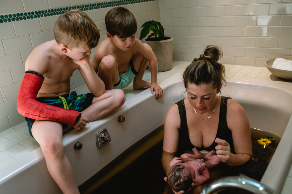 Mother lifting newborn from warm herbal bath while siblings watch and support nearby