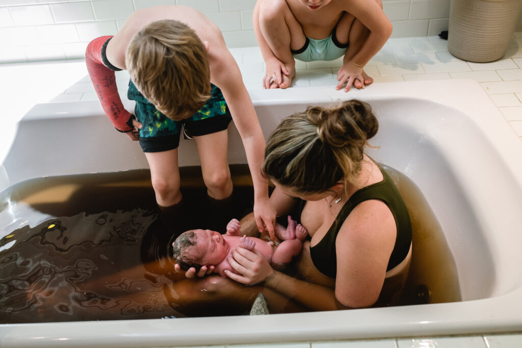 Children leaning over bathtub watching newborn float in herbal bath after home birth