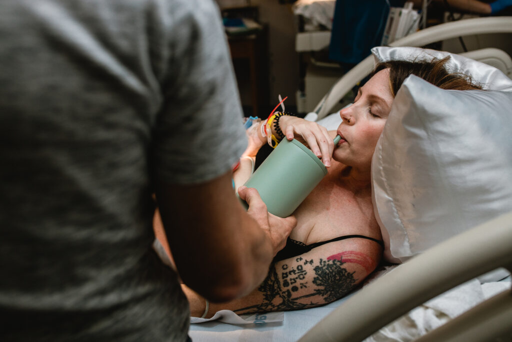 Mother sipping water through a straw while laboring in a hospital bed, supported by her husband nearby
