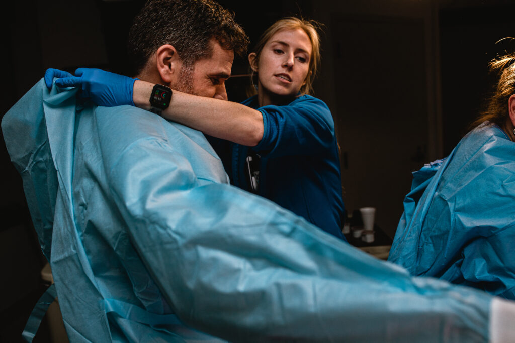 Labor and delivery nurse helping father into sterile gown as he prepares to assist with catching the baby