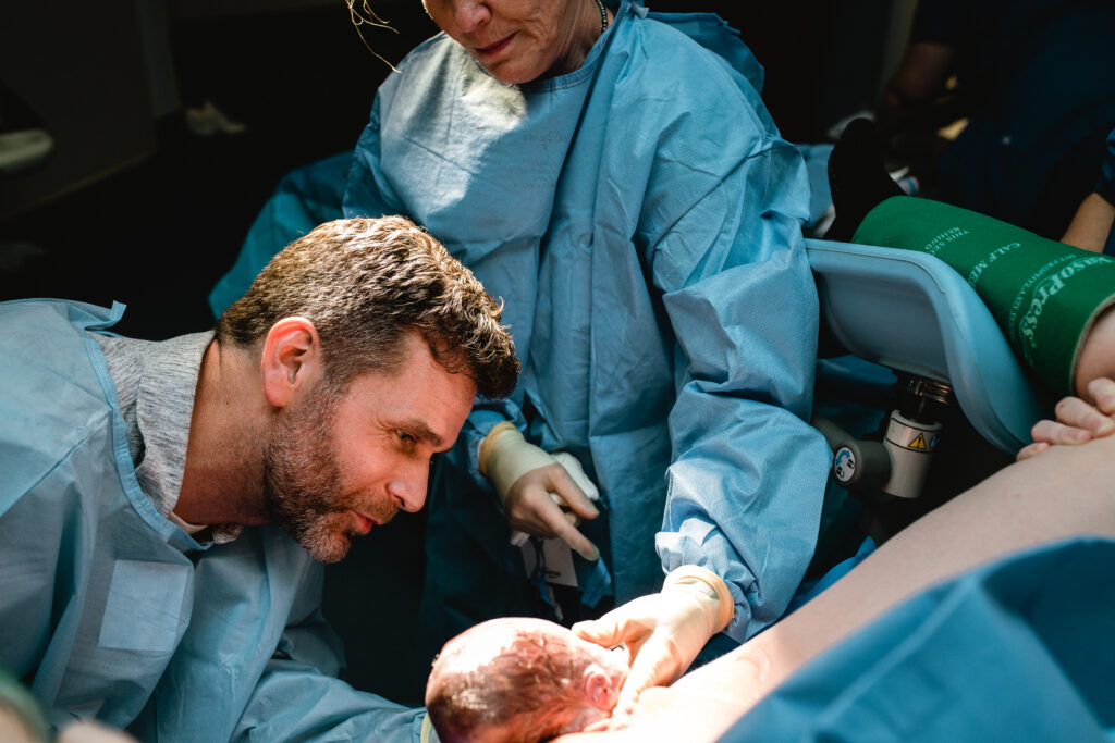 Father assisting the OB at the foot of the bed during the final moments before baby is born
