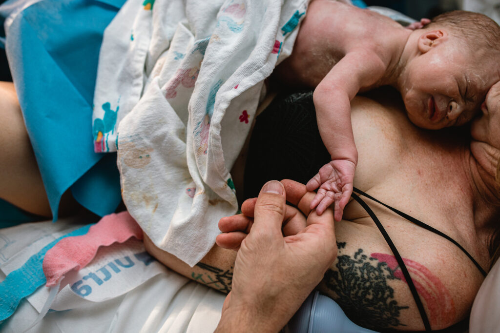 Newborn baby lying on mother’s chest after birth while father gently holds the baby’s tiny hand during golden hour hospital delivery
