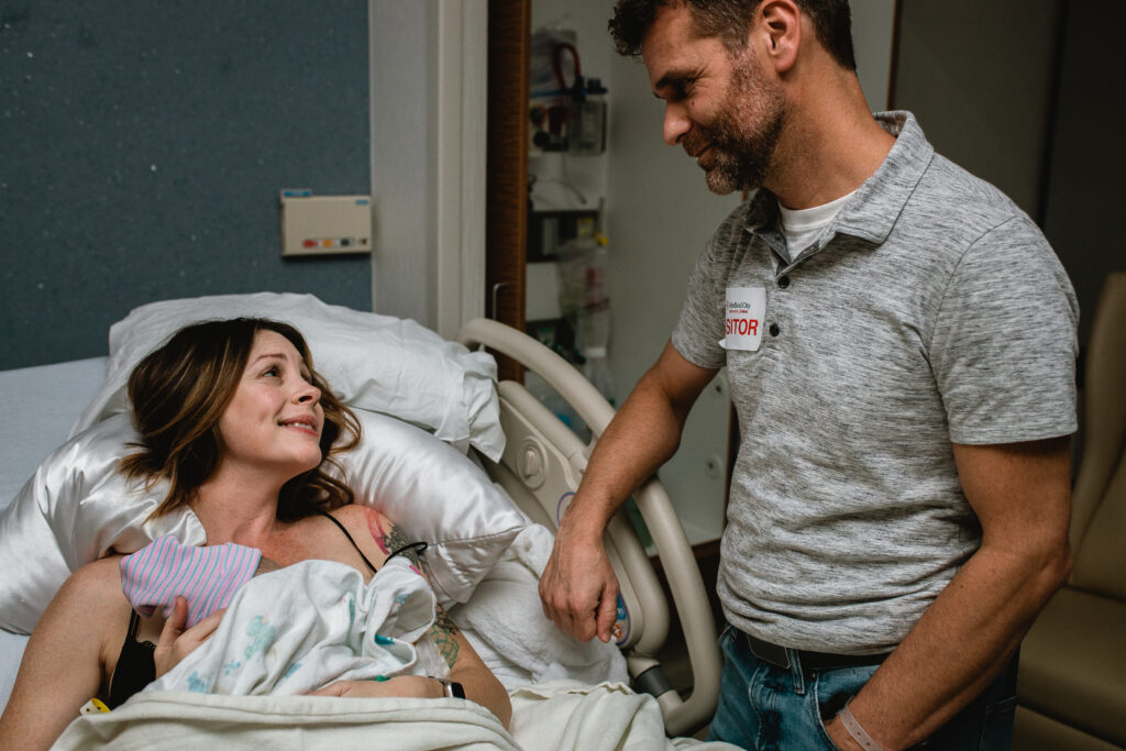 Mother resting in hospital bed with newborn on her chest and OB nurse nearby providing postpartum care